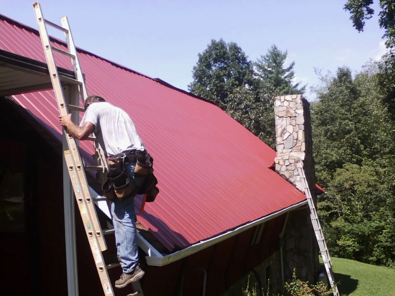 Roofer on a ladder beside a red metal roof and stone chimney — Soffit & Fascia Repair by Roofing Brothers in Kimberton